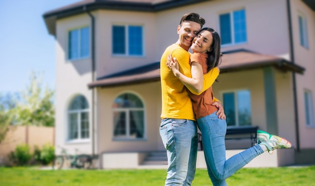 Foto moderna hermosa pareja joven emocionada enamorada de pie frente a la nueva casa grande con las llaves en las manos y abrazándose mientras celebran esta compra