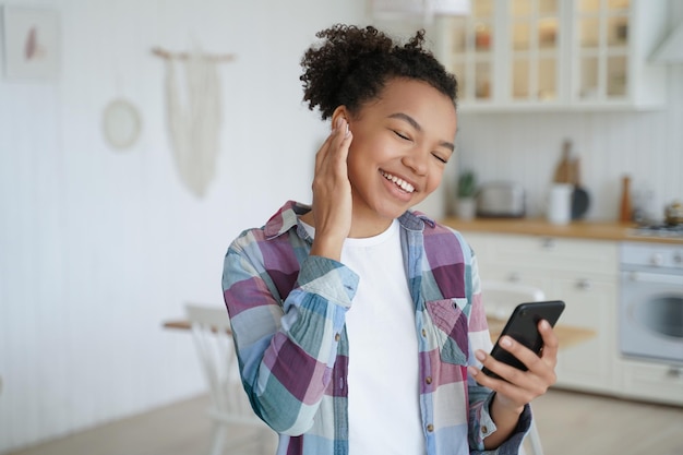 Moderna joven mestiza escuchando música sosteniendo un teléfono inteligente usando aplicaciones musicales en casa