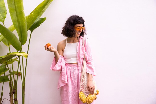 Moderna joven morena caucásica mira hacia un lado sosteniendo una bolsa de hilo con mandarinas sobre fondo blanco Concepto de feliz fin de semana