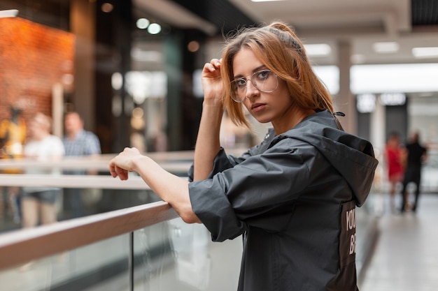 Moderna joven pelirroja con elegante chaqueta con capucha en gafas de moda está de pie en una tienda cerca de la pared de cristal brillante. Chica de moda disfruta de un paseo por el centro comercial. Estilo.