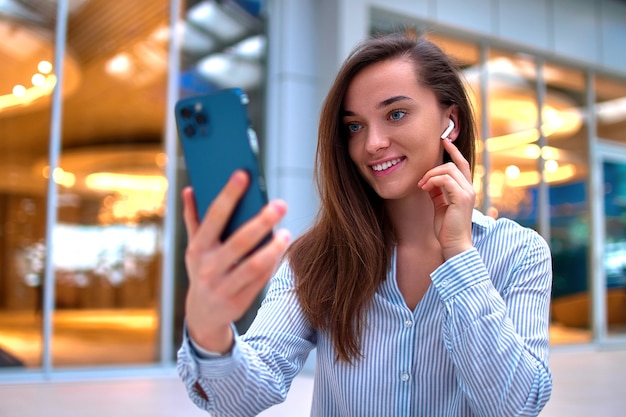 Moderna mujer milenaria inteligente casual feliz usando auriculares inalámbricos usando un teléfono inteligente para videollamadas y chatear en línea de forma remota