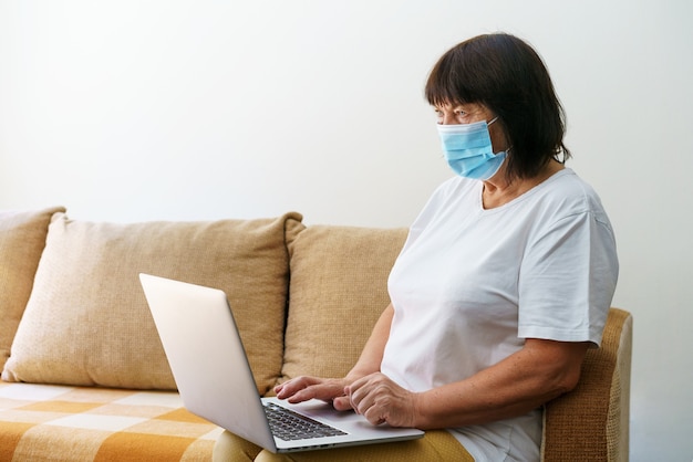 Moderne ältere Frau mit medizinischer Maske, die zu Hause auf dem Sofa sitzt und Computer-Gadget verwendet, glückliche Reife ...