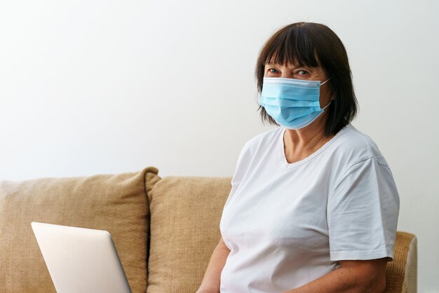 Moderne ältere Frau mit medizinischer Maske, die zu Hause auf dem Sofa sitzt und Computer-Gadget verwendet, glückliche Reife ...