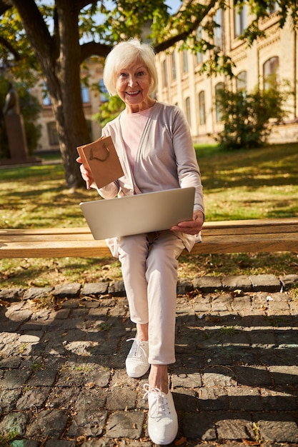 moderne, alternde Frau, die glücklich lächelt, während sie einen Laptop in einer Hand und eine Mini-Tasche in der anderen hält