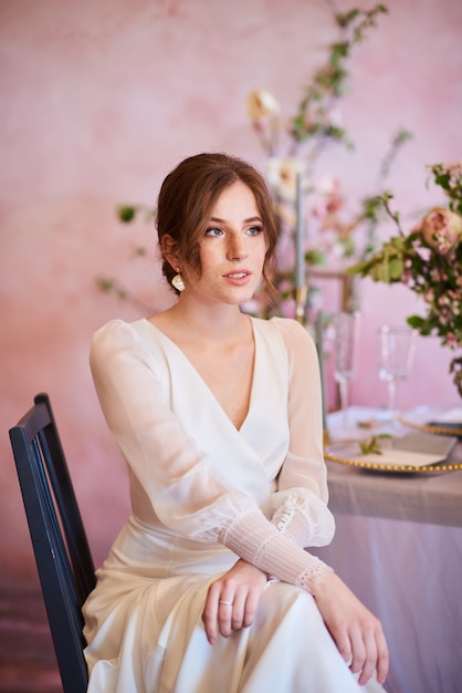 Moderne Braut in einem schönen weißen Kleid. Zarte Braut mit Sommersprossen sitzt nachdenklich an einem Hochzeitstisch im Bankettbereich.