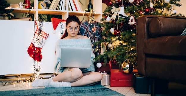 moderne Frau, die auf dem Laptop tippt, während sie neben geschmücktem Weihnachtsbaum und leichten Girlanden sitzt