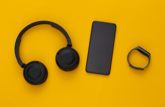 Moderne Geräte. Smartphone mit schwarzen Stereo-Kopfhörern und einem Smart-Armband auf einem gelben