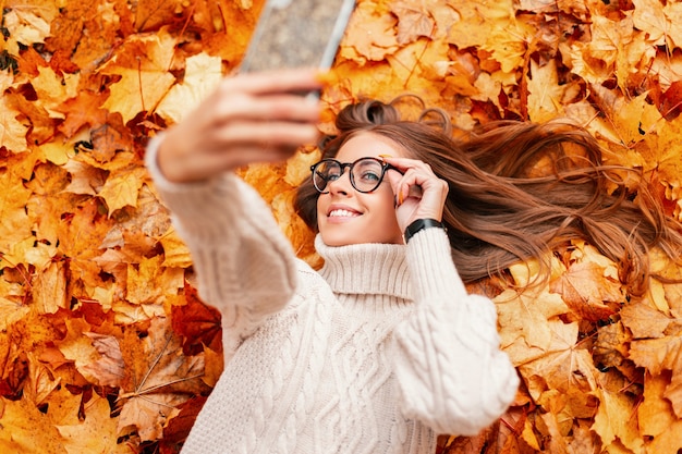Moderne glückliche junge Hipsterfrau in stilvollen Gläsern in einem modischen Strickpullover liegt auf orangefarbenen Blättern im Park und macht ein Selfie auf einem Telefon. Trendy schönes Mädchen lächelnd. Sicht von oben.