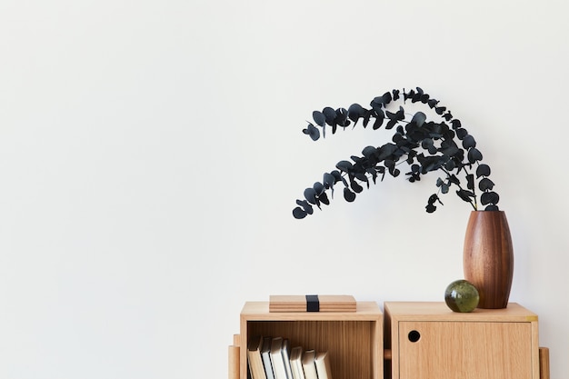 Moderne Komposition des Wohnzimmers mit Design-Bücherregal aus Holz, Eukalyptusblatt in Vase, Buch, Dekoration, Glaskugel und Kopienraum an der weißen Wand