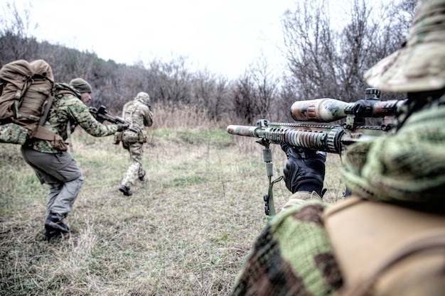 Moderne Kriegsführungsgruppe, Kommandokämpfer-Trupp, Teammitglieder, die angreifen, durch Wälder eilen. Scharfschütze oder Scharfschütze, die Kameraden im Wald bedeckt, über Schulteransicht