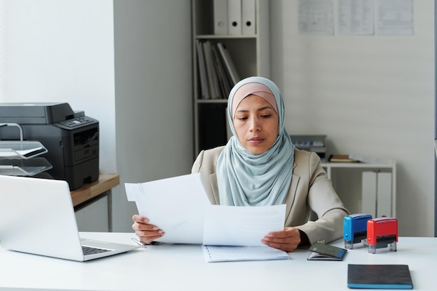 Moderne muslimische Frau mit Hijab, die am Schreibtisch im Büro sitzt und Papierkram erledigt, um Informationen zu überprüfen