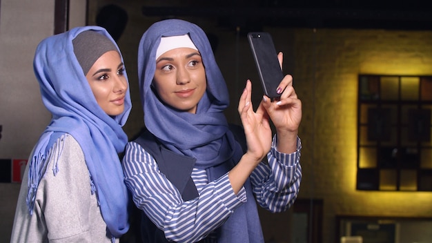 Moderne muslimische Frauen fotografieren auf einem Handy. Mädchen in Hijabs reden und lächeln.