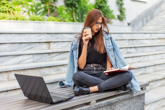 Moderne Studenten. Fernunterricht. Junge begeisterte Frau liest Buch, während auf Bank mit einem Laptop sitzend