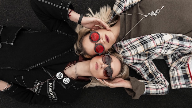 Moderne urbane, stilvolle junge Schwestern in trendigen Brillen in modischer Jugendkleidung, die auf der Straße ruht. Frisches Porträt zwei sexy schöne Freundinnen Blondinen auf der Straße