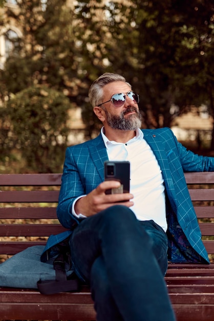 Moderner älterer Geschäftsmann, der Smartphone für Online-Meetings verwendet, während er auf einer Bank sitzt.