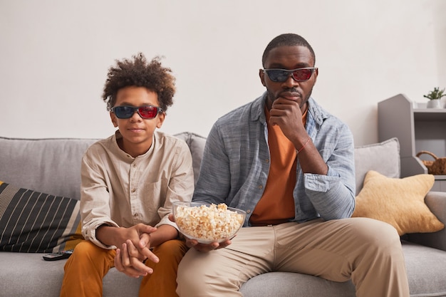 Moderner afroamerikanischer Familienvater und sein jugendlicher Sohn sitzen auf dem Sofa im Wohnzimmer, schauen sich einen 3D-Film an und essen Popcorn