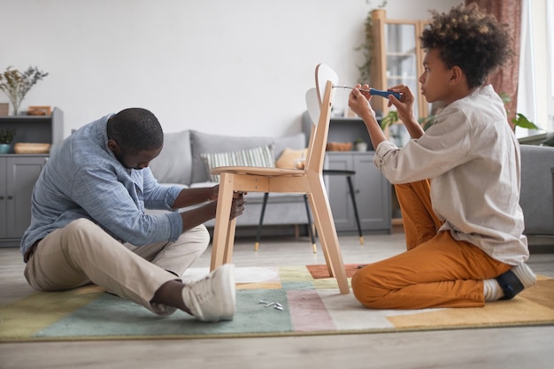 Moderner afroamerikanischer Mann verbringt Zeit mit seinem jugendlichen Sohn im Wohnzimmer zu Hause und baut einen neuen Stuhl zusammen