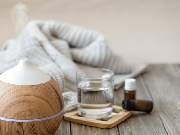 Moderner Aromaöldiffusor auf Holzoberfläche mit Strickelement, Wasser und Ölen in Gläsern.