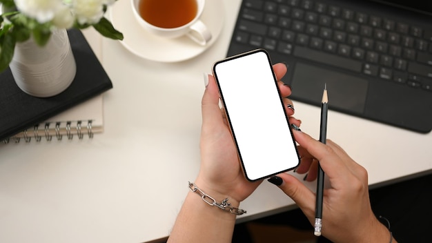 Moderner Büroarbeitsplatz mit weiblichen Händen, die eine Draufsicht des weißen Bildschirmmodells des Smartphones halten