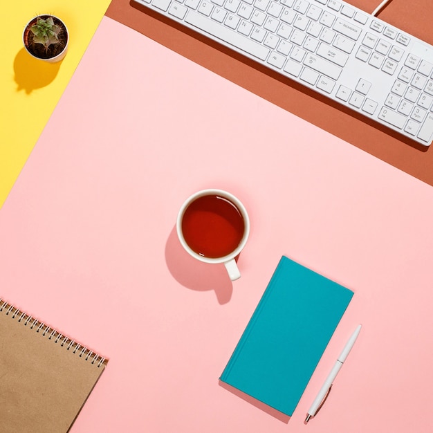 Moderner bunter Freiberuflerarbeitsplatz. Flache Zusammensetzung von Tastatur, Kaktus, Tagebuch mit Stift und Tasse Tee auf buntem Schreibtisch. Rosa, Gelb, Aquamarin und und braune Farben