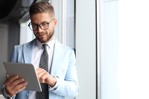 Moderner Geschäftsmann in formeller Kleidung mit digitalem Tablet, während er in der Nähe des Fensters im Büro steht.