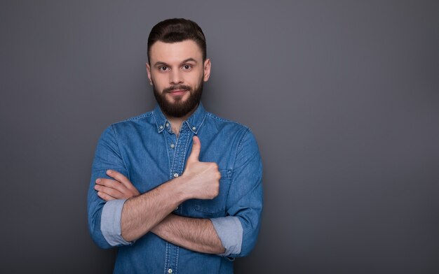 Moderner glücklicher aufgeregter junger bärtiger Mann im Jeanshemd zeigt Daumen nach vorne und hat Spaß