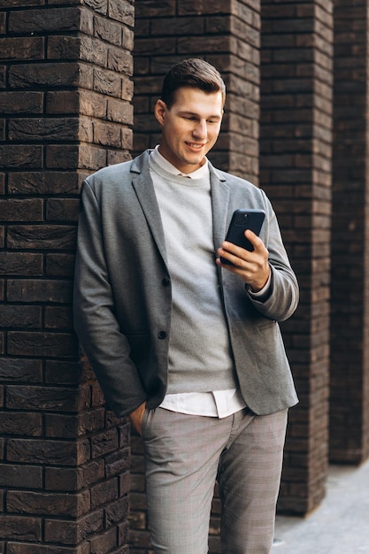 Moderner gutaussehender lächelnder Mann, der an der Wand auf der Straße steht und ein Telefon hält