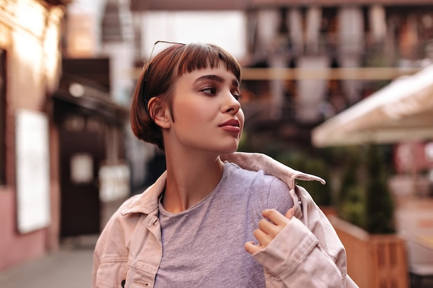 Moderner Hipster mit kurzen Haaren in hellem Outfit, der draußen posiert Modische brünette Dame in rosa Jeansjacke, die auf die Straße blickt