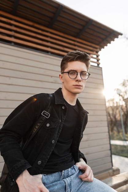 Moderner junger Mann in schwarzer Jeansjacke in blauer Vintage-Jeans mit Brille sitzt in der Nähe eines Holzgebäudes auf der Straße