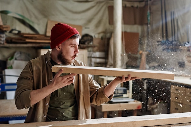 Moderner männlicher Tischler mit roter Strickmütze, der in der Schreinerei Holzbretter hält, die Staub davon blasen