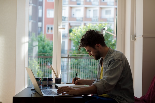 moderner Mann des hübschen Hippies, der nach Hause unter Verwendung des Laptops arbeitet