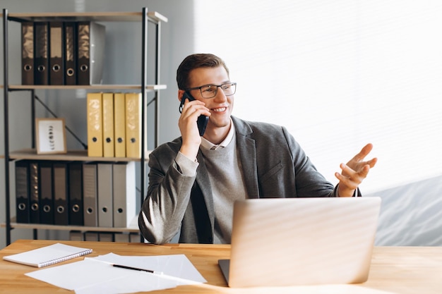 Moderner Mann mit Brille, lächelnder Büroangestellter, der am Laptop arbeitet und ein Telefongespräch führt