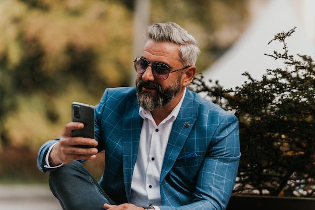 Moderner Senior-Geschäftsmann nutzt Smartphone für Online-Meetings, während er auf einer Bank sitzt.