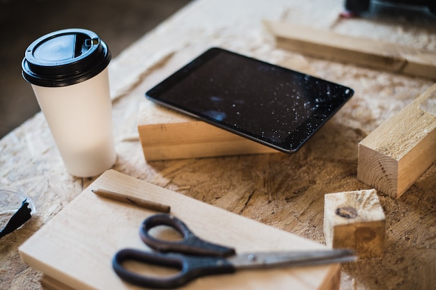 Moderner Tischlerarbeitsplatz mit Tablette, Kaffee, Schere und Holzstücken auf Tisch, in Werkstatt