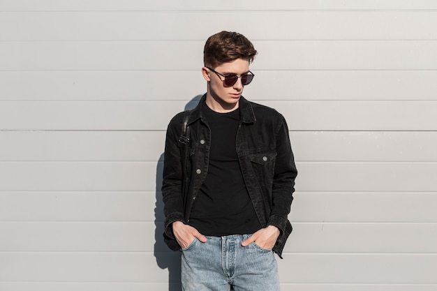 Moderner trendiger junger Mann in modischer Jeanskleidung in stylischer Sonnenbrille