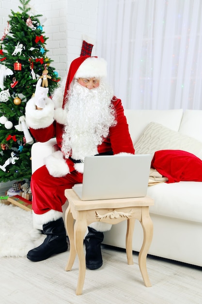 Moderner Weihnachtsmann, der auf Sofa sitzt und Computer benutzt