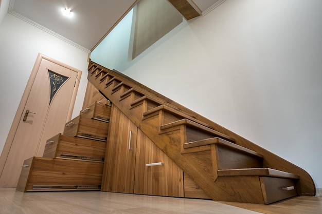 Modernes Architekturinterieur mit luxuriösem Flur mit glänzender Holztreppe in einem mehrstöckigen Haus Maßgeschneiderte ausziehbare Schränke auf Gleitern in Schlitzen unter der Treppe Nutzung von Stauraum