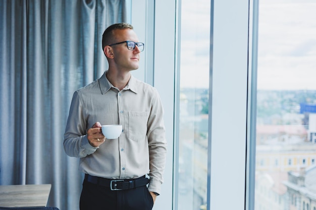 Modernes Büro Kaufmann oder Manager trinkt Kaffee am Fenster lächelnd Porträt eines erfolgreichen IT-Ingenieur-Programmierers mit Brille, der im Büro arbeitet