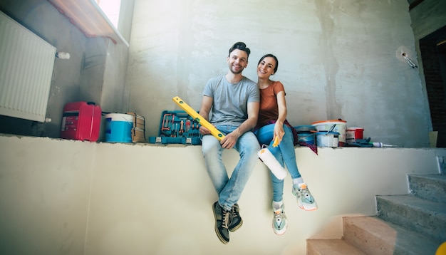 Modernes glückliches junges Paar verliebt in Freizeitkleidung während der Reparatur oder Renovierung von Wänden mit vielen Werkzeugen dafür