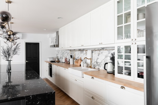 Modernes klassisches Kücheninterieur mit Küchengeräten und weißem Keramikspülbecken mit goldenem Spiegelhahn auf Holzplatte mit Marmorwand
