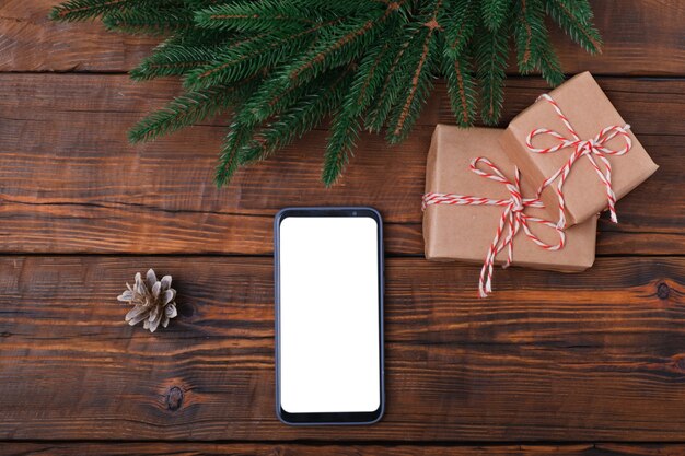 Modernes Smartphone mit Modell auf Vintage Holztisch mit Weihnachtsdekoration, Geschenkbox. Flache Lay-Vorlage. Tannenkranz