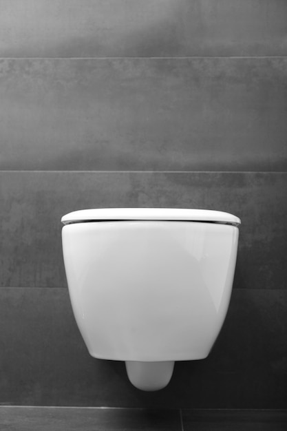 Un moderno inodoro blanco montado en la pared con tapa cerrada y un botón de descarga cromado brillante contra una pared negra Inodoro en el interior del baño