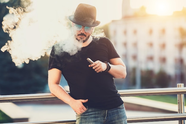 Moderno joven feliz con sombrero con barba divertido Vaporizadores En el fondo la puesta de sol de la tarde