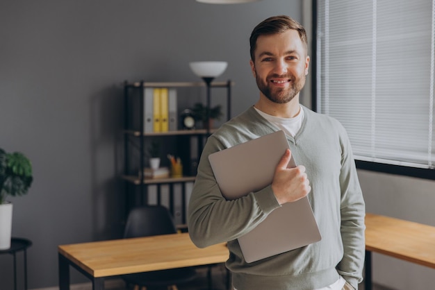 Moderno trabalhador de escritório barbudo feliz de uma empresa de TI sorrindo e segurando um laptop no escritório