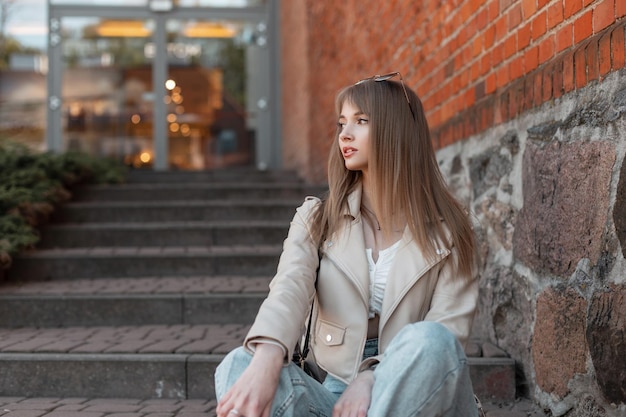 Modeschönes junges Zoomer-Mädchen in modischer Freizeitkleidung mit stylischer Lederjacke und Jeans sitzt auf den Stufen in der Nähe der Ziegelwand eines Vintage-Einkaufszentrums