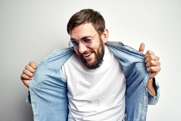 Modisch aussehender kaukasischer, gutaussehender sexy Mann mit stylischer Brille, cool posierend und gestikulierend, weißes T-Shirt unter seinem Hemd zeigend, emotional ausdrucksstarkes Gesicht isoliert im Studio