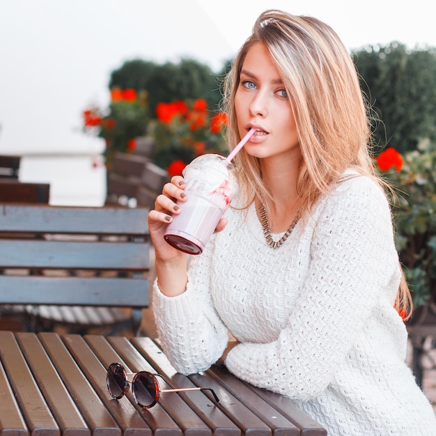 Modisch schönes junges Mädchen in einem weißen Pullover sitzt an einem Sommertag und trinkt einen Cocktail