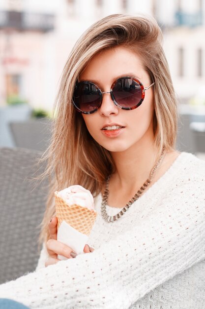 Modische attraktive junge Hipster-Frau in einem gestrickten stilvollen Pullover in der trendigen Sonnenbrille mit Eis in den Händen sitzt in einem Café an einem sonnigen Frühlingstag. Nettes Mädchen blond.