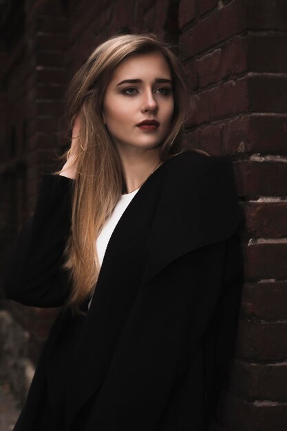 Modische blonde Frau im warmen Mantel nahe einer Backsteinmauer