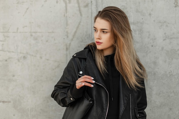 Modische entzückende Frau Modell in Mode Rock Leder schwarze Kleidung mit Lederjacke schwarzen Hoodie und Leder schwarze Tasche steht in der Nähe einer grauen Betonwand Urbanes weibliches Porträt